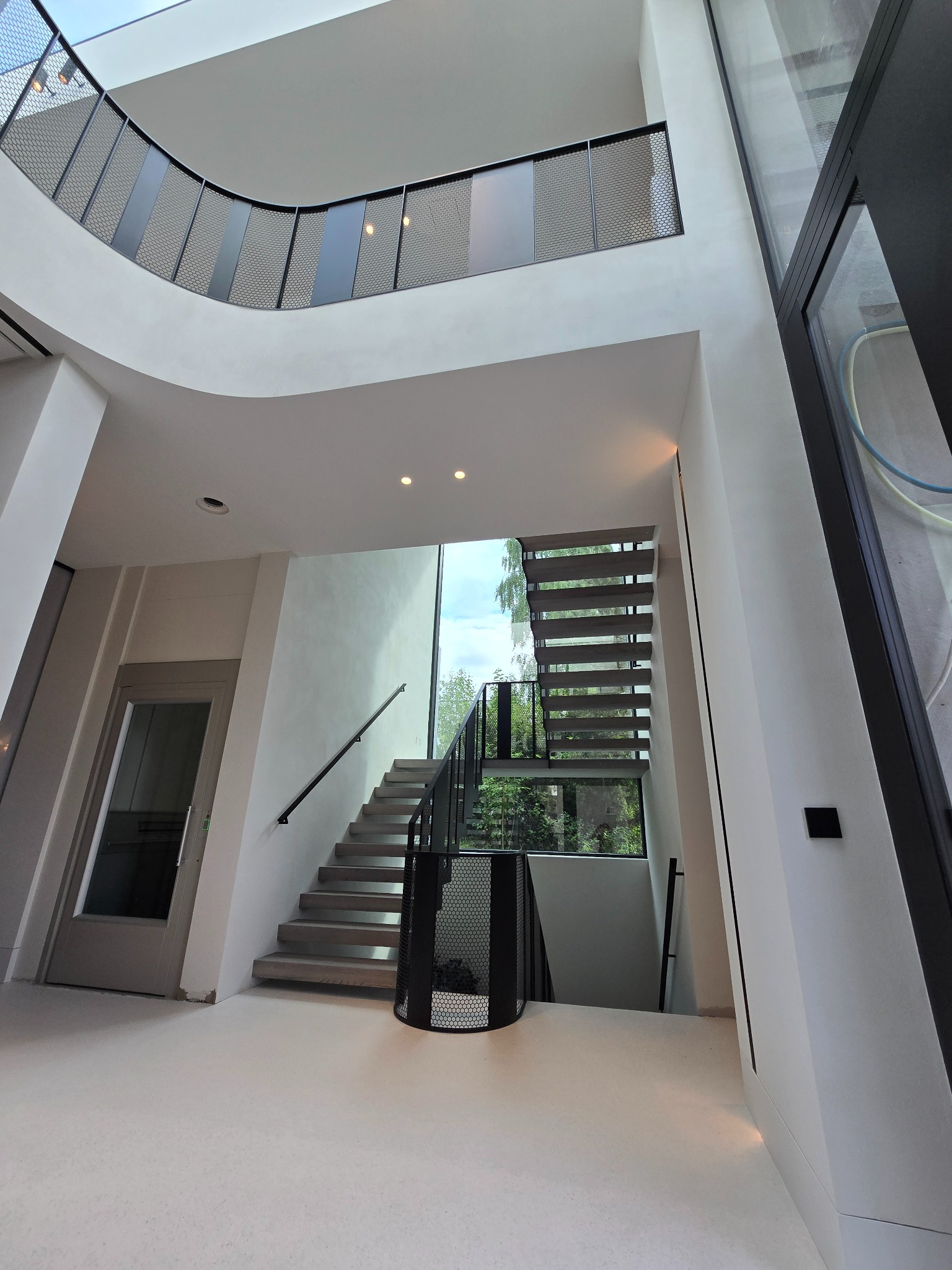 Wnętrze nowoczesnego domu z wysokim atrium, schodami z drewnianymi stopniami i czarną, ażurową balustradą, widok z dołu na antresolę z podobną balustradą, duże okna wpuszczają naturalne światło.