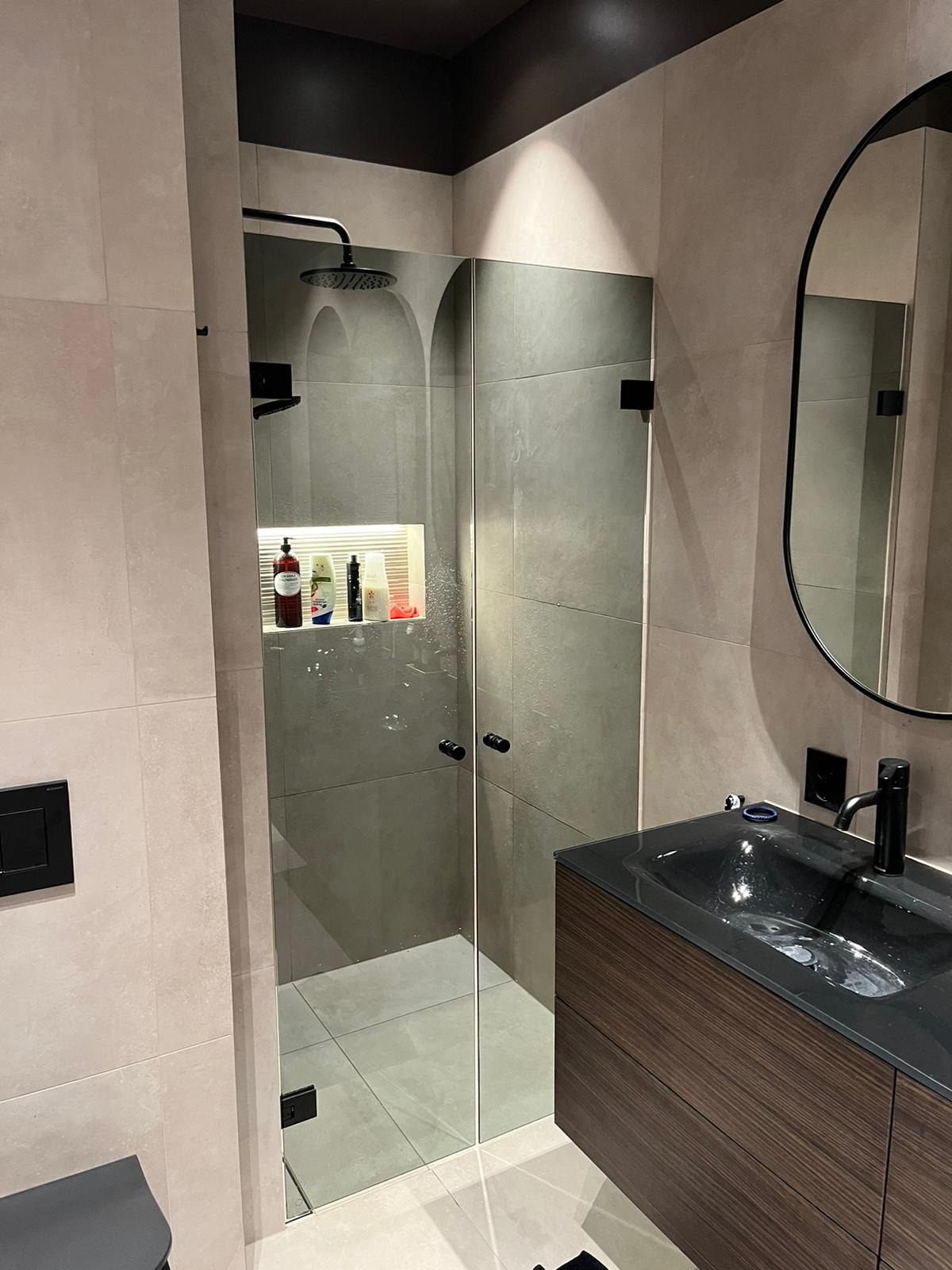 Nowoczesna łazienka z kabiną prysznicową typu walk-in, szklane drzwi z czarnymi zawiasami, umywalka z szafką w kolorze ciemnego drewna i okrągłe lustro.