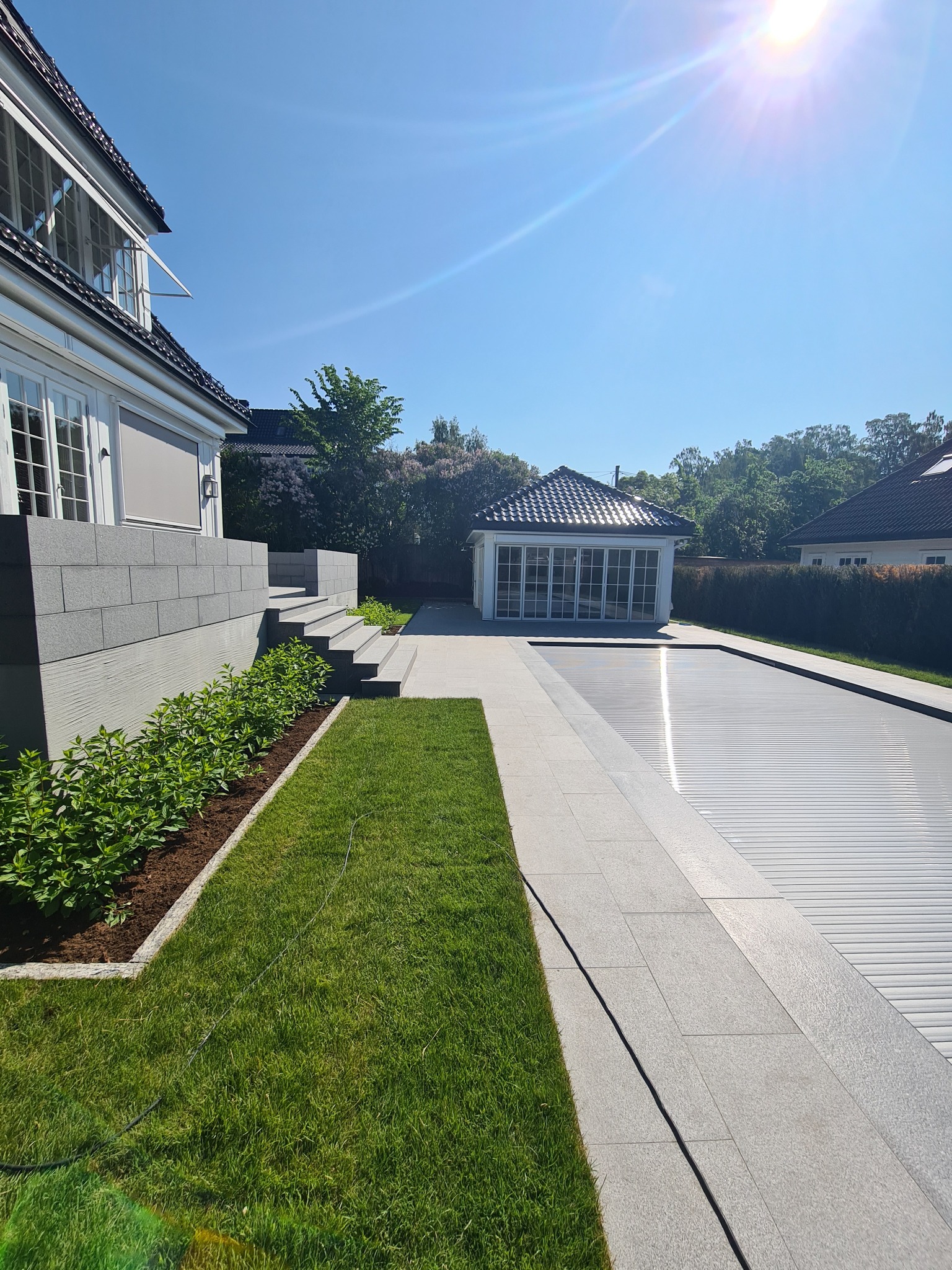 Nowoczesny dom z szarymi kamiennymi schodami i tarasem prowadzącym do zadaszonego basenu z roletą, oświetlony jasnym słońcem.