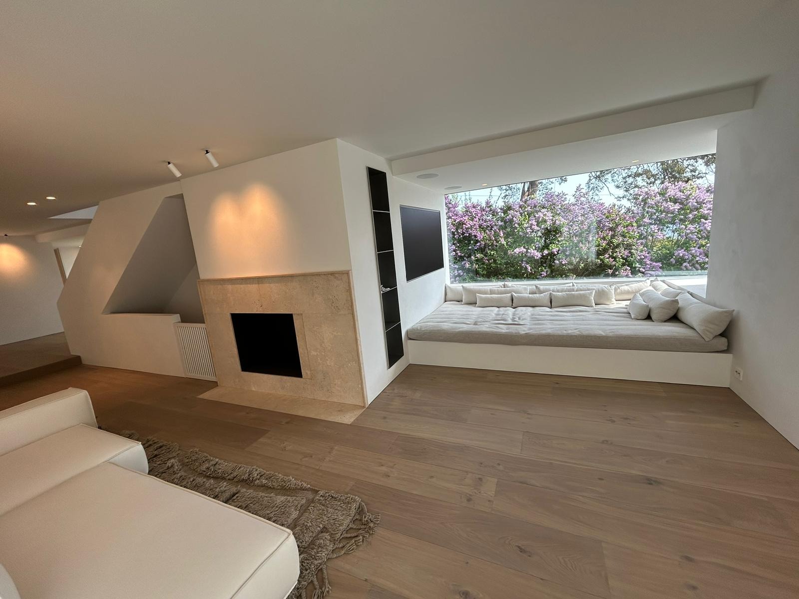 Jasne wnętrze z kominkiem obłożonym beżowym kamieniem i wnęką okienną z siedziskiem i poduszkami, za oknem kwitnące krzewy.