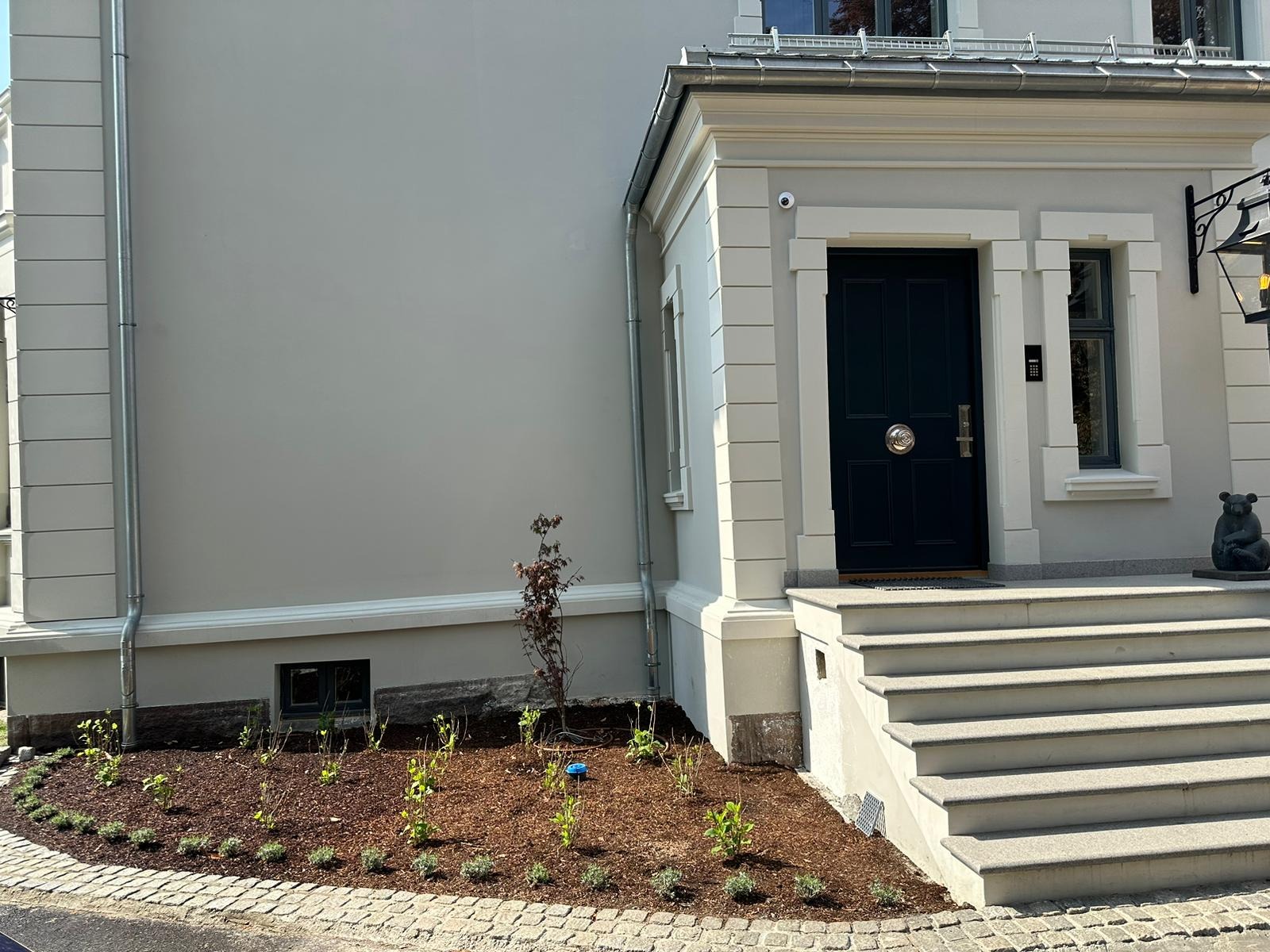 Elewacja domu w jasnych odcieniach szarości z ciemnymi drzwiami wejściowymi i schodami, świeżo założona rabata z korą i młodymi roślinami, widoczny fragment chodnika z kostki brukowej.