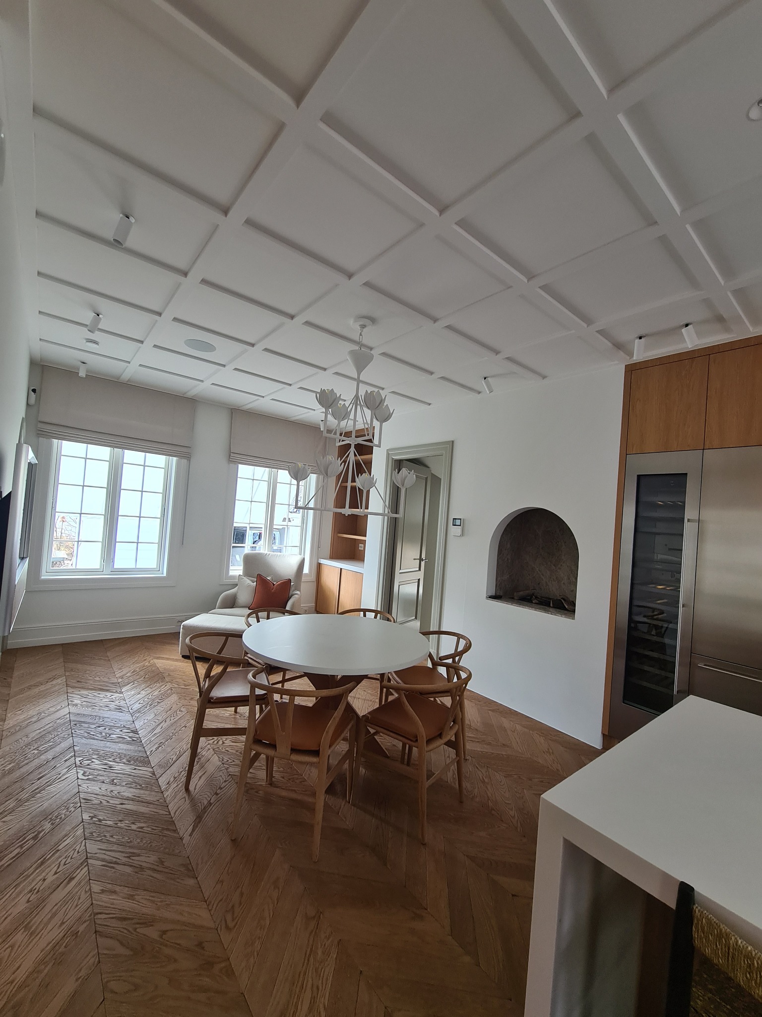 Jasne wnętrze z parkietem w jodełkę, okrągłym stołem z drewnianymi krzesłami, designerskim żyrandolem i dekoracyjnym sufitem kasetonowym.