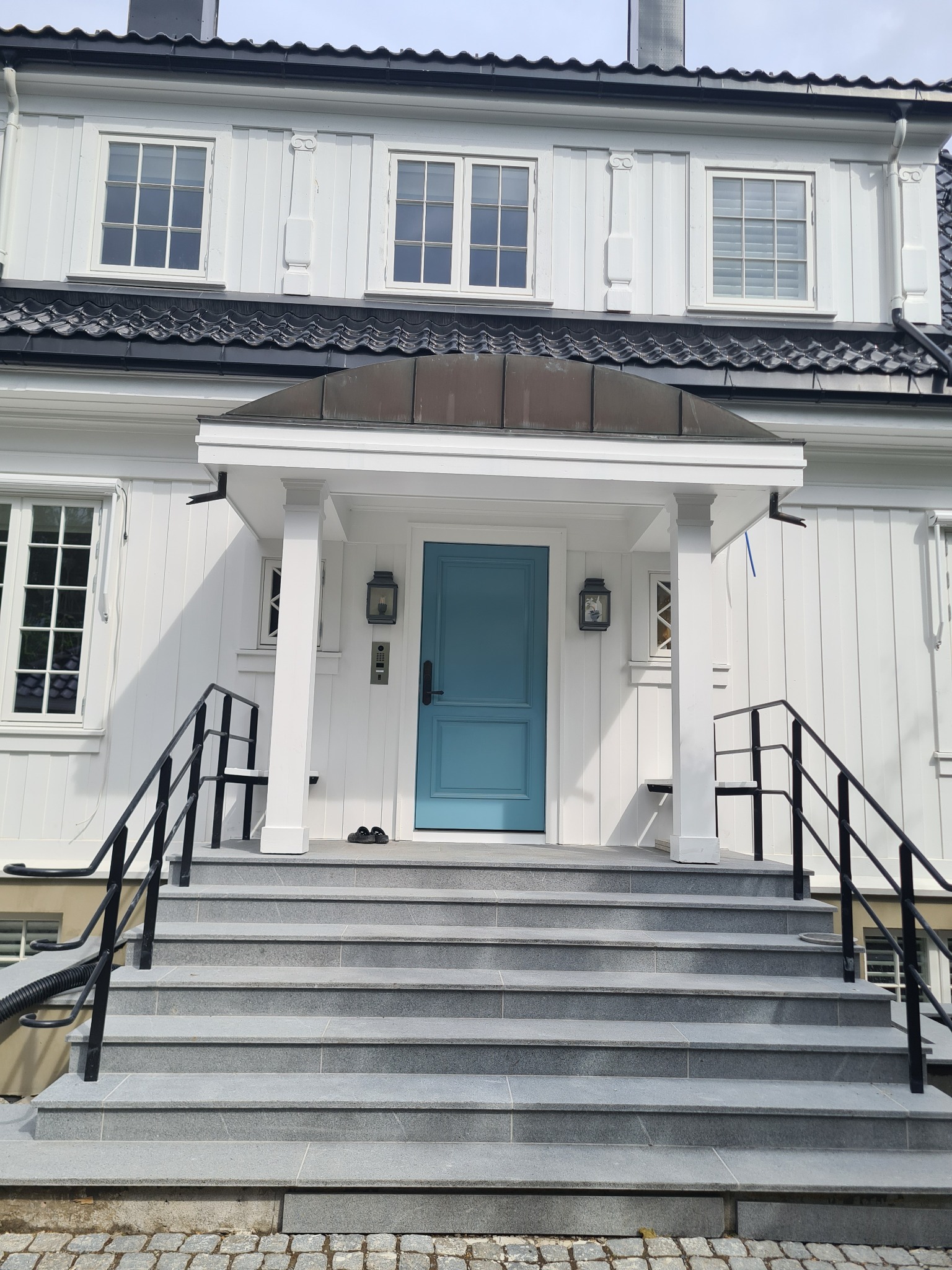 Elewacja domu z jasnymi deskami, niebieskimi drzwiami wejściowymi i szarymi schodami z czarną balustradą.