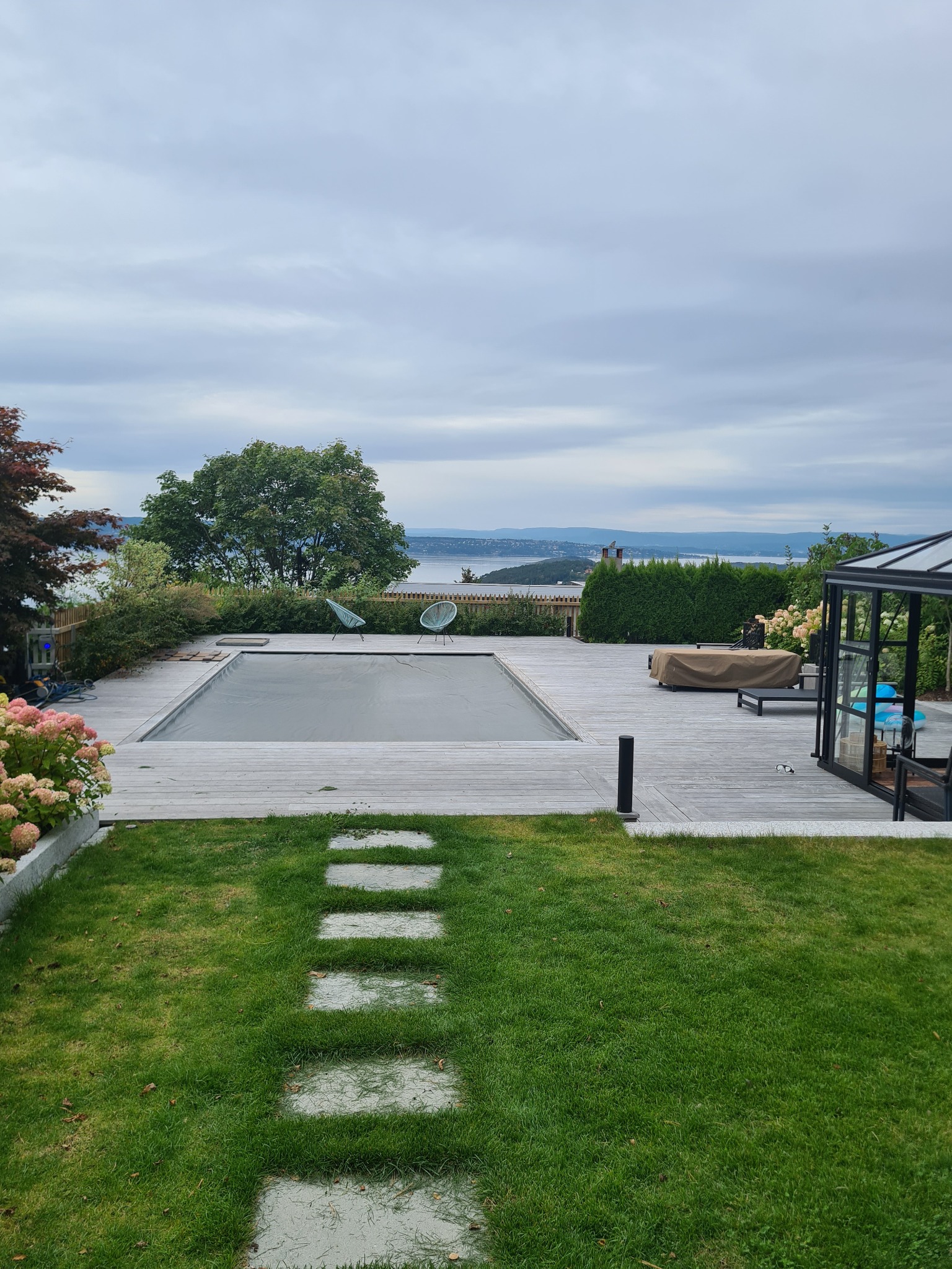 Widok na basen z przykryciem, otoczony drewnianym tarasem i zielonym trawnikiem z kamienną ścieżką, w tle krajobraz jeziora i gór pod pochmurnym niebem.