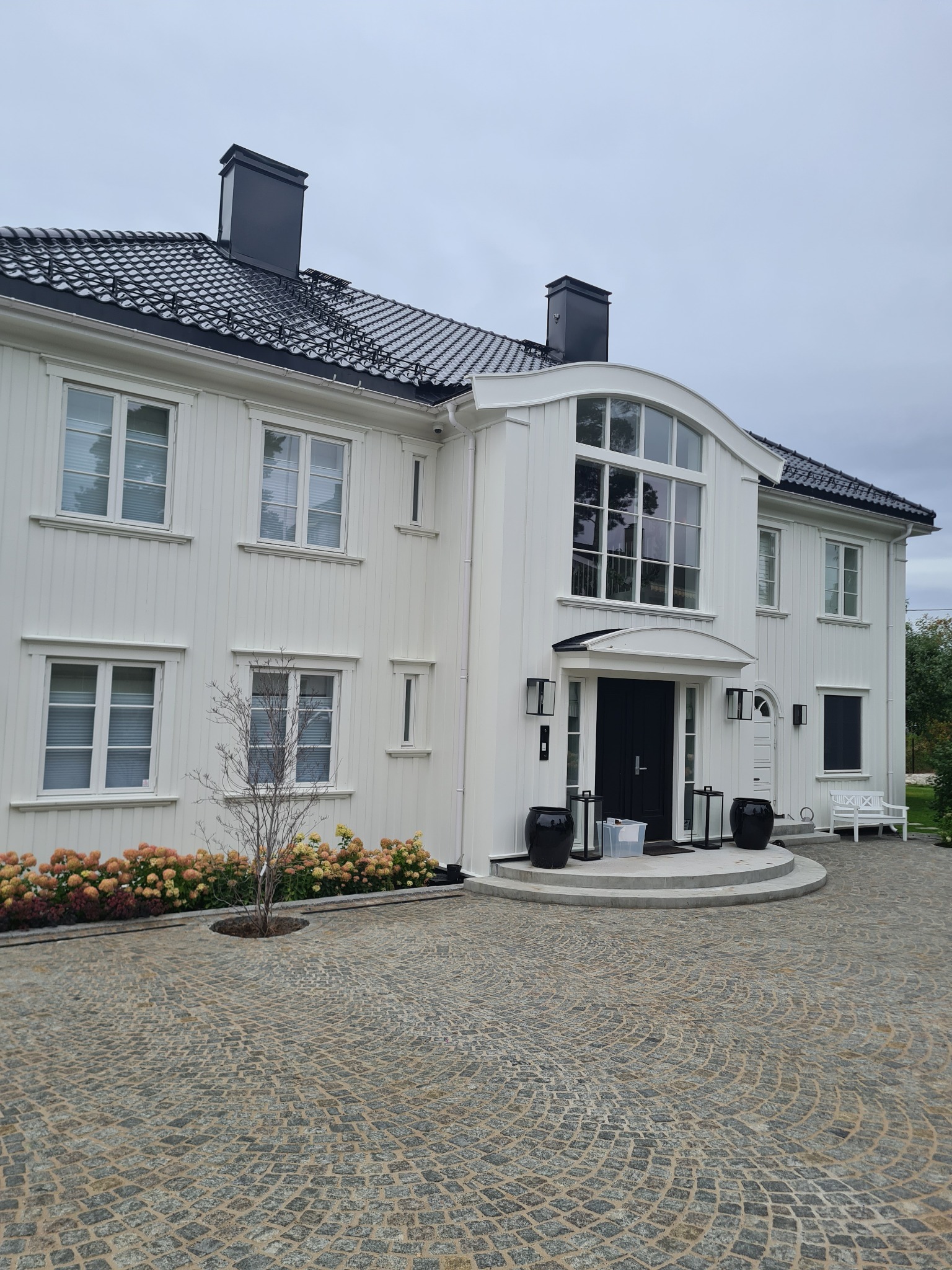 Elegancki, dwukondygnacyjny dom z białymi panelami, czarnym dachem i czarnymi akcentami, takimi jak komin i donice, z brukowanym podjazdem i rabatą z hortensjami.