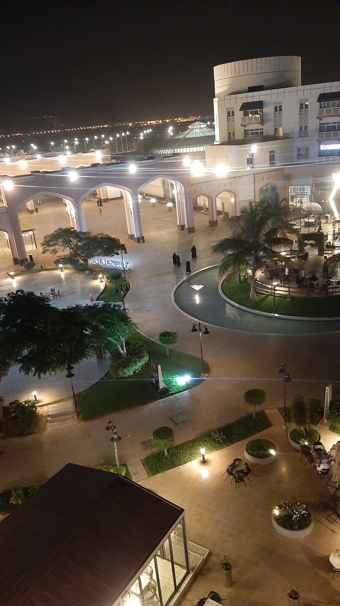 Nocny widok z góry na oświetlony plac miejski z palmami, fontanną i budynkami w stylu arabskim.