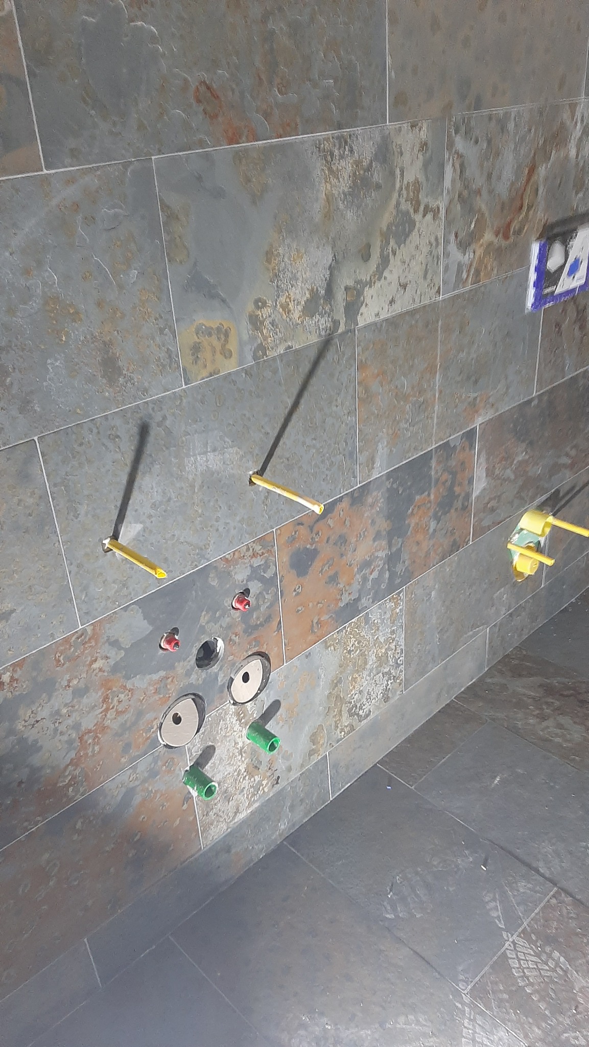 Ściana w łazience wyłożona płytkami imitującymi łupek, z widocznymi instalacjami wodno-kanalizacyjnymi w trakcie montażu, w tym wystające rury i otwory na baterie.