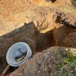 PTSBUD-Expert - Wykopany dół z betonowym kręgiem i rurą kanalizacyjną, widoczny fragment koparki. Przygotowanie do instalacji szamba lub oczyszczalni.