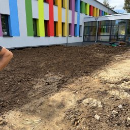 PTSBUD-Expert Tomasz Strzałka - Przygotowany teren pod nasadzenia przed budynkiem z kolorowymi pasami okiennymi. Widoczna ziemia, częściowo ułożona kostka brukowa, narzędzia ogrodnicze w tle.