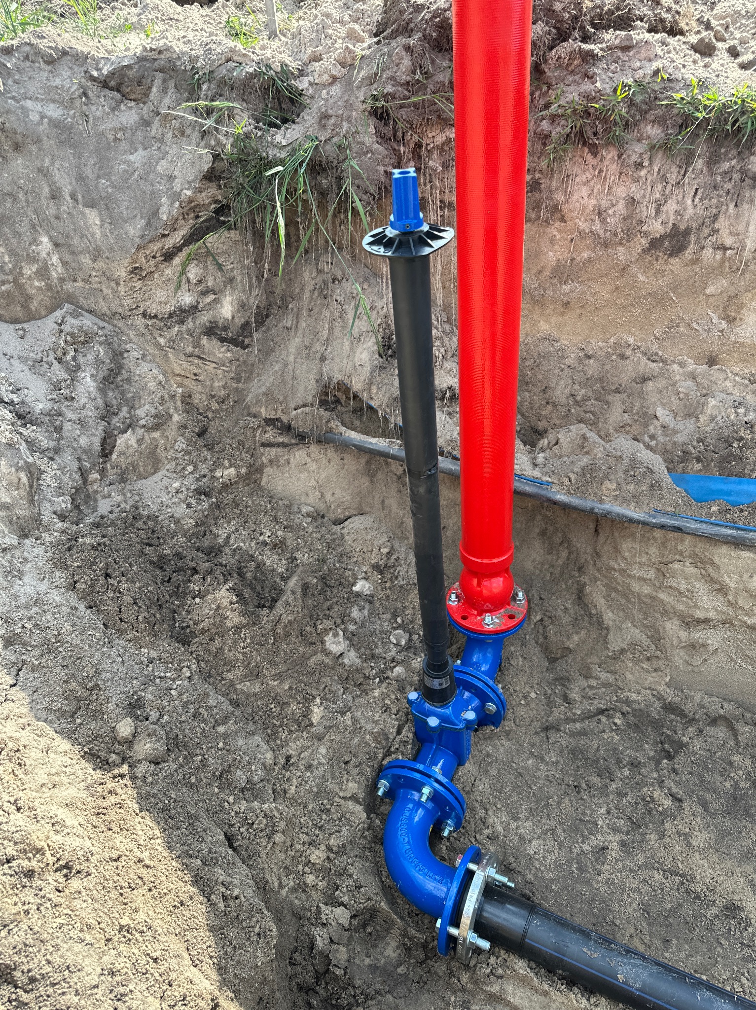 Niebieskie złącze rur z kołnierzami, czerwona i czarna rura w wykopie ziemnym. Widoczne inne rury. Instalacja wodociągowa w trakcie prac ziemnych.