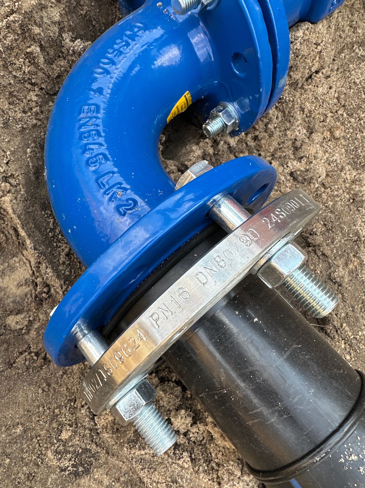 Niebieski kolanko z kołnierzem i czarna rura połączone śrubami na piasku. Widoczne oznaczenia DN80/90 i EN616 LK2. Element instalacji wodociągowej w trakcie montażu.