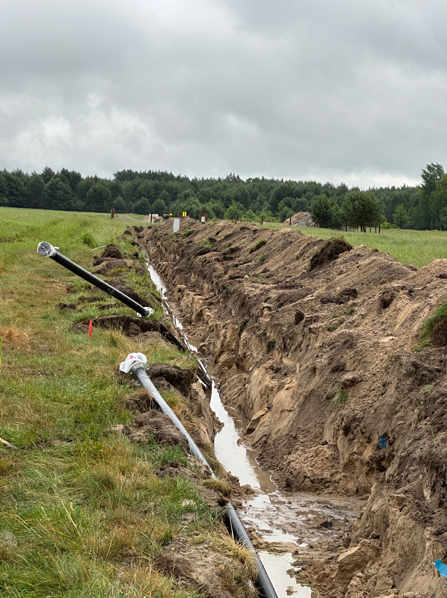 Wykopany rów wypełniony wodą, z wystającymi rurami z tworzywa sztucznego na zewnątrz, na tle zielonego pola i lasu pod pochmurnym niebem. Widoczne ślady prac ziemnych.