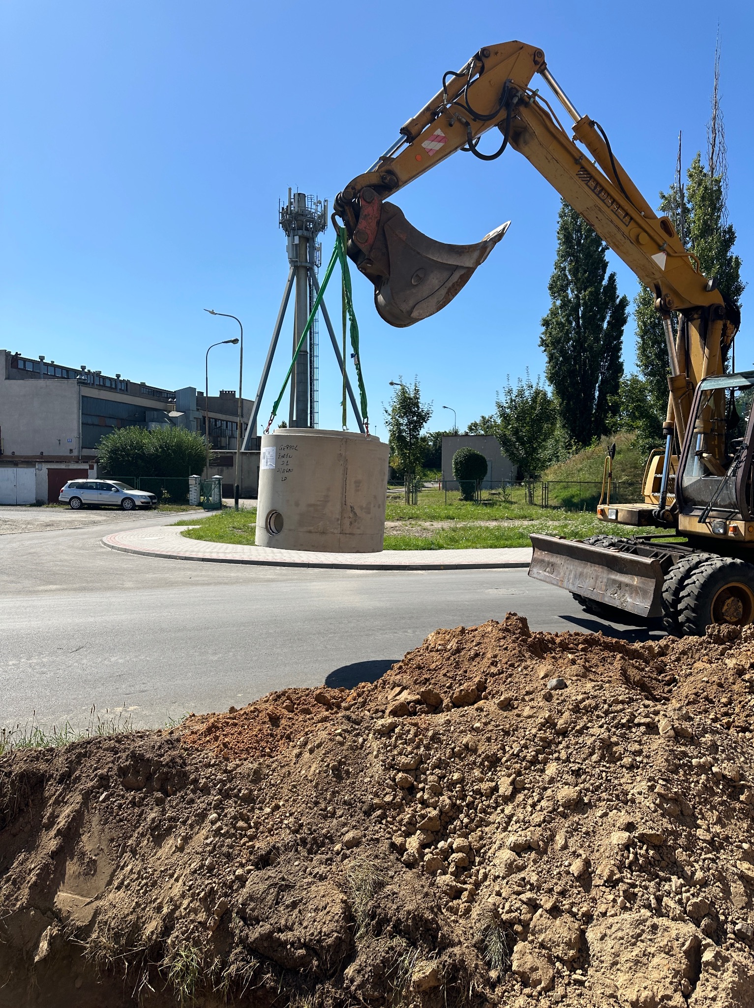 Żółta koparka umieszcza betonowy zbiornik w wykopie na tle miejskiego krajobrazu i błękitnego nieba. Widoczne wykopana ziemia i fragment ulicy.