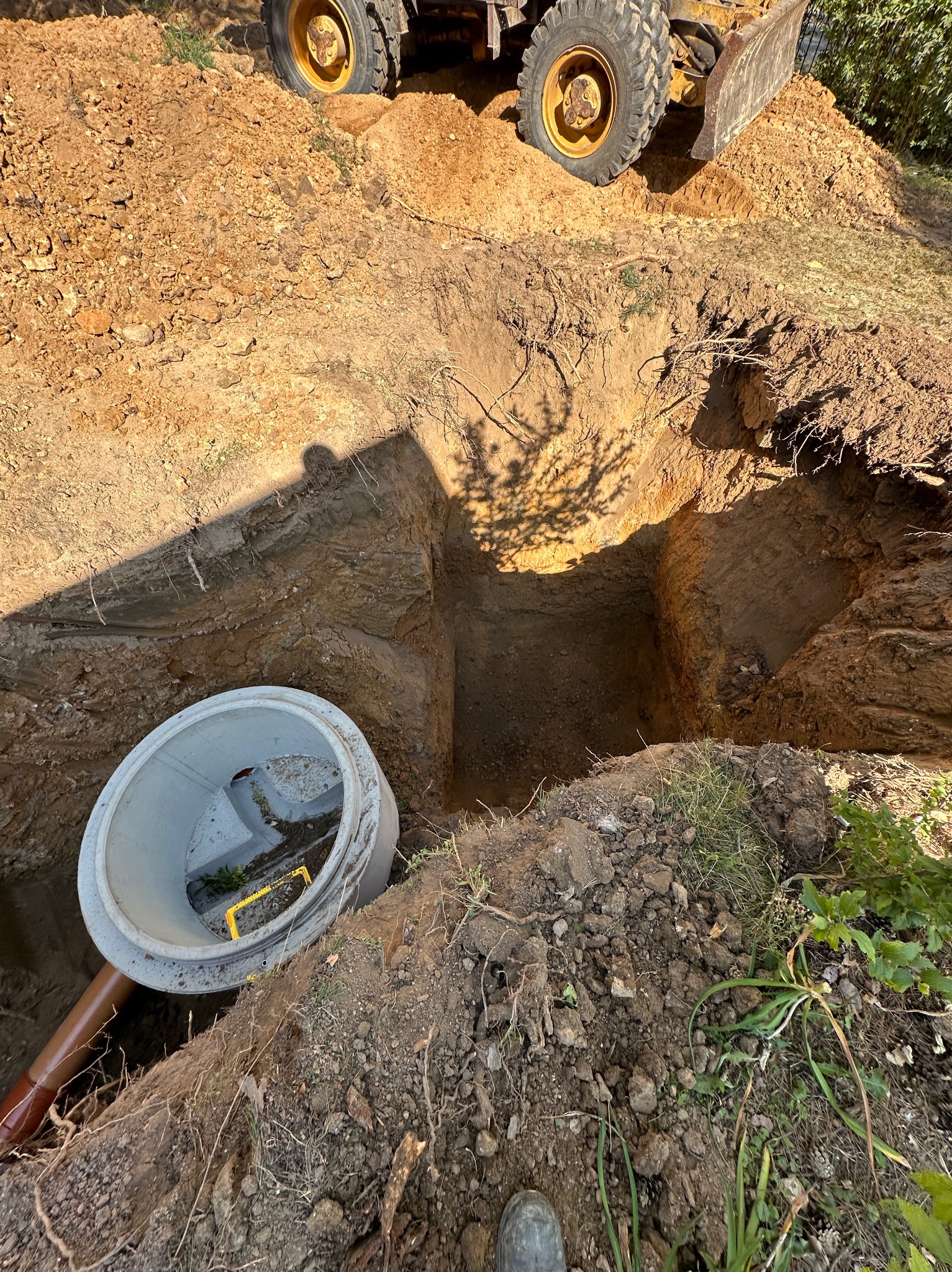 Wykopany dół z betonowym kręgiem i rurą kanalizacyjną, widoczny fragment koparki. Przygotowanie do instalacji szamba lub oczyszczalni.