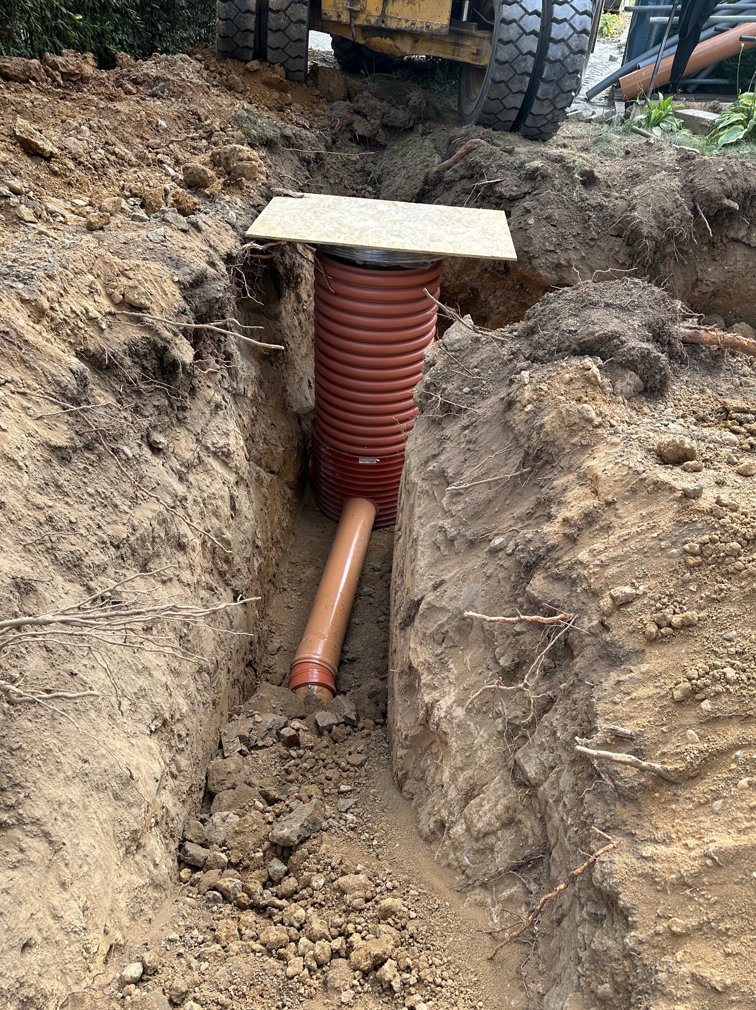 Wykop w ziemi z pomarańczową rurą kanalizacyjną i studzienką z pokrywą. W tle widoczne koła koparki. Prace ziemne przy instalacji kanalizacji.