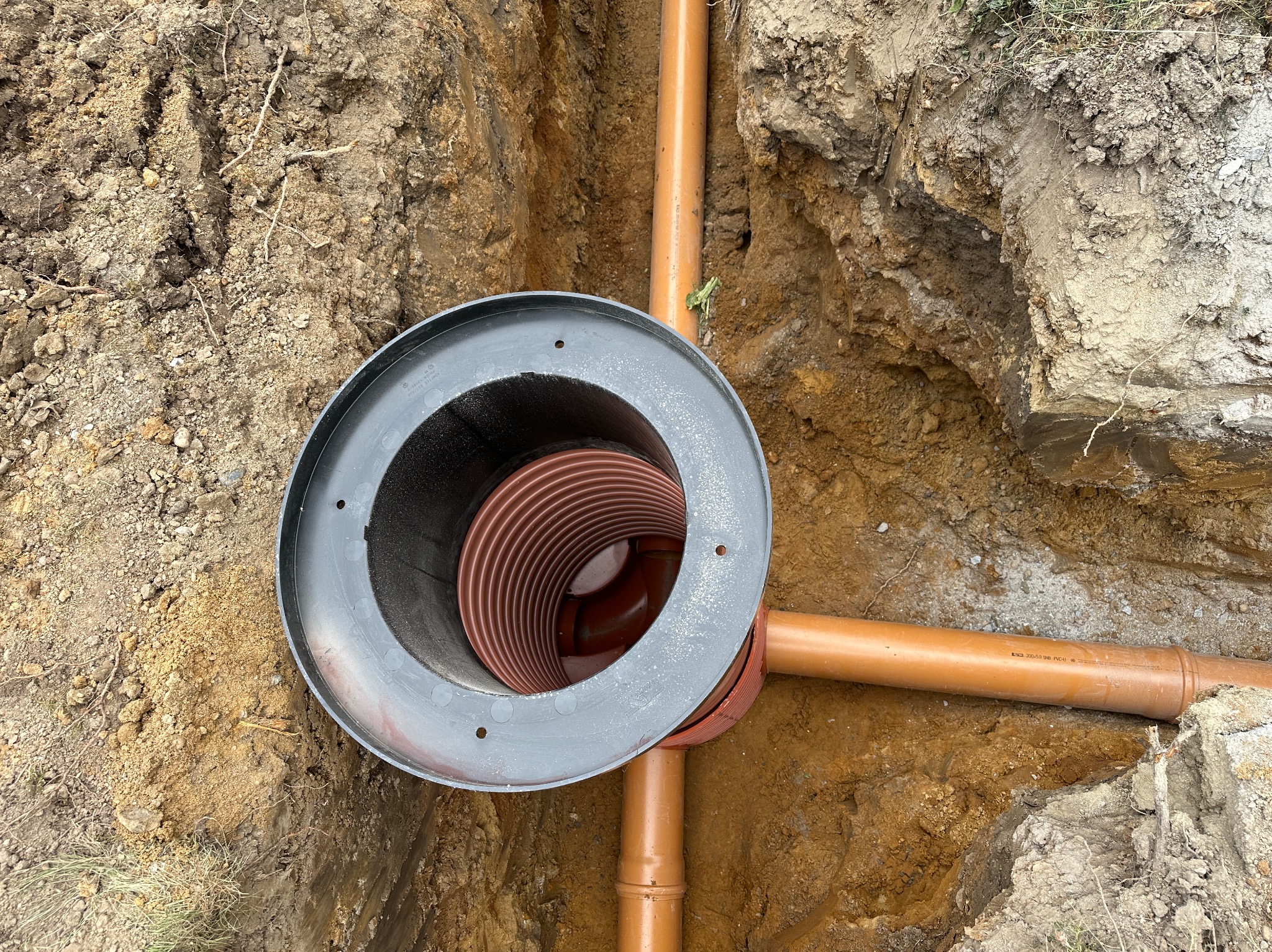 Studzienka kanalizacyjna z brązowymi rurami w wykopie ziemnym. Widok z góry na instalację w trakcie budowy, z wyraźnymi śladami wykopu.