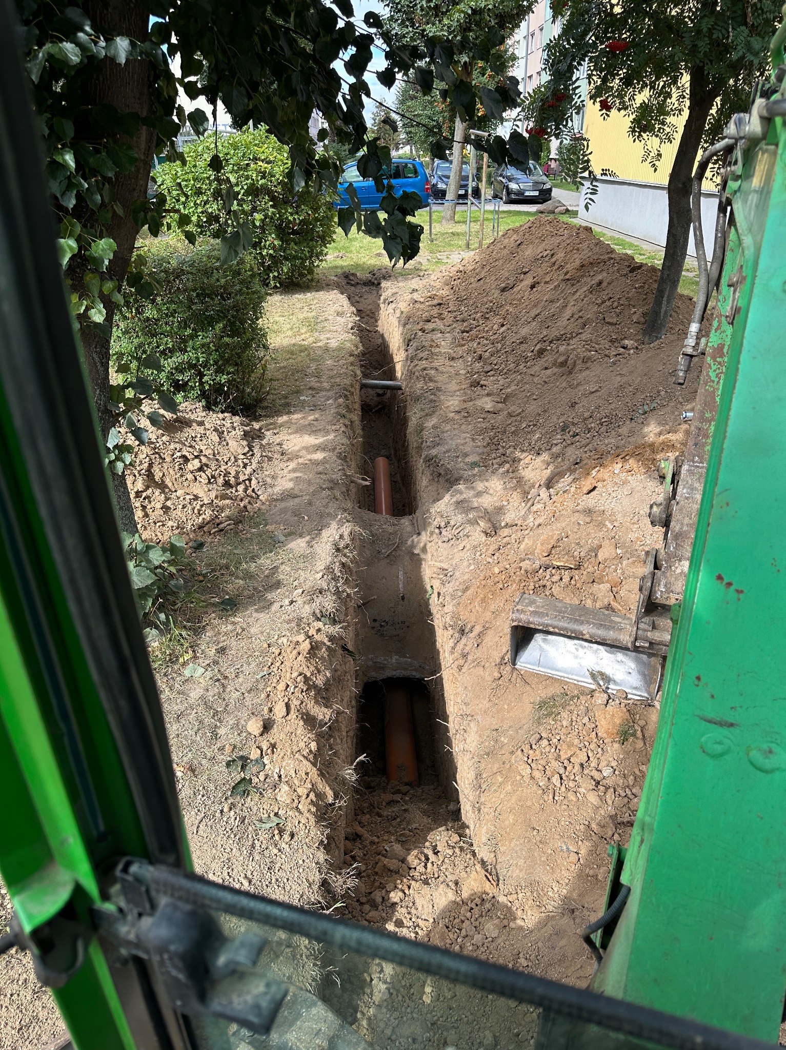 Wykopany wąski rów z pomarańczowymi rurami na dnie, widok z kabiny zielonej koparki, obok hałda ziemi, drzewa i budynki w tle. Prace ziemne w przestrzeni miejskiej.
