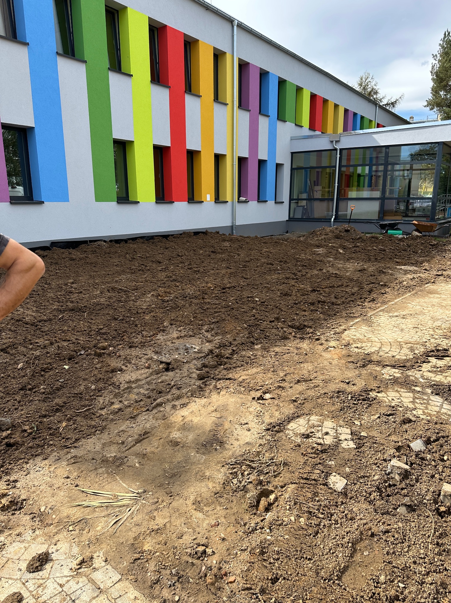 Przygotowany teren pod nasadzenia przed budynkiem z kolorowymi pasami okiennymi. Widoczna ziemia, częściowo ułożona kostka brukowa, narzędzia ogrodnicze w tle.