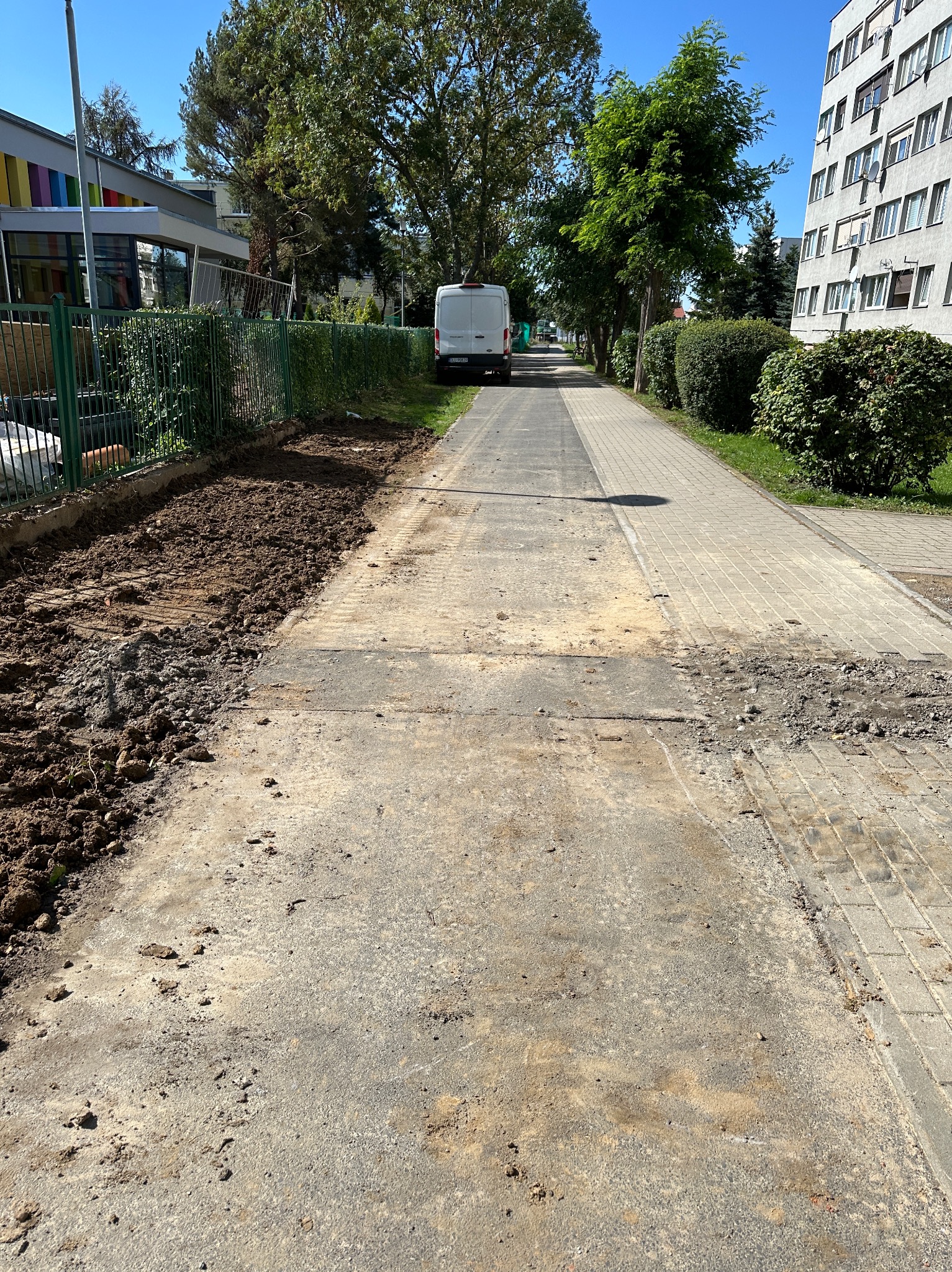 Przygotowanie terenu pod chodnik: wykopana ziemia wzdłuż ogrodzenia, widoczne ślady po pracach ziemnych, w tle budynki mieszkalne i biały samochód dostawczy.