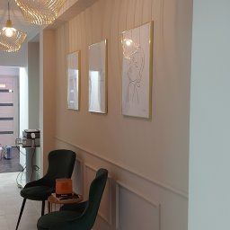 Krystian Ciuruś KRYST-REM - Jasne wnętrze poczekalni z kremowymi ścianami ozdobionymi sztukaterią i trzema obrazami w złotych ramkach, oświetlone dekoracyjnymi lampami wiszącymi, z dwoma ciemnozielonymi krzesłami i stolikiem...
