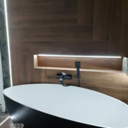 Nowoczesna łazienka z wolnostojącą czarną wanną, drewnopodobnymi płytkami na ścianie z podświetlaną półką i szarą ścianą z oświetleniem LED.