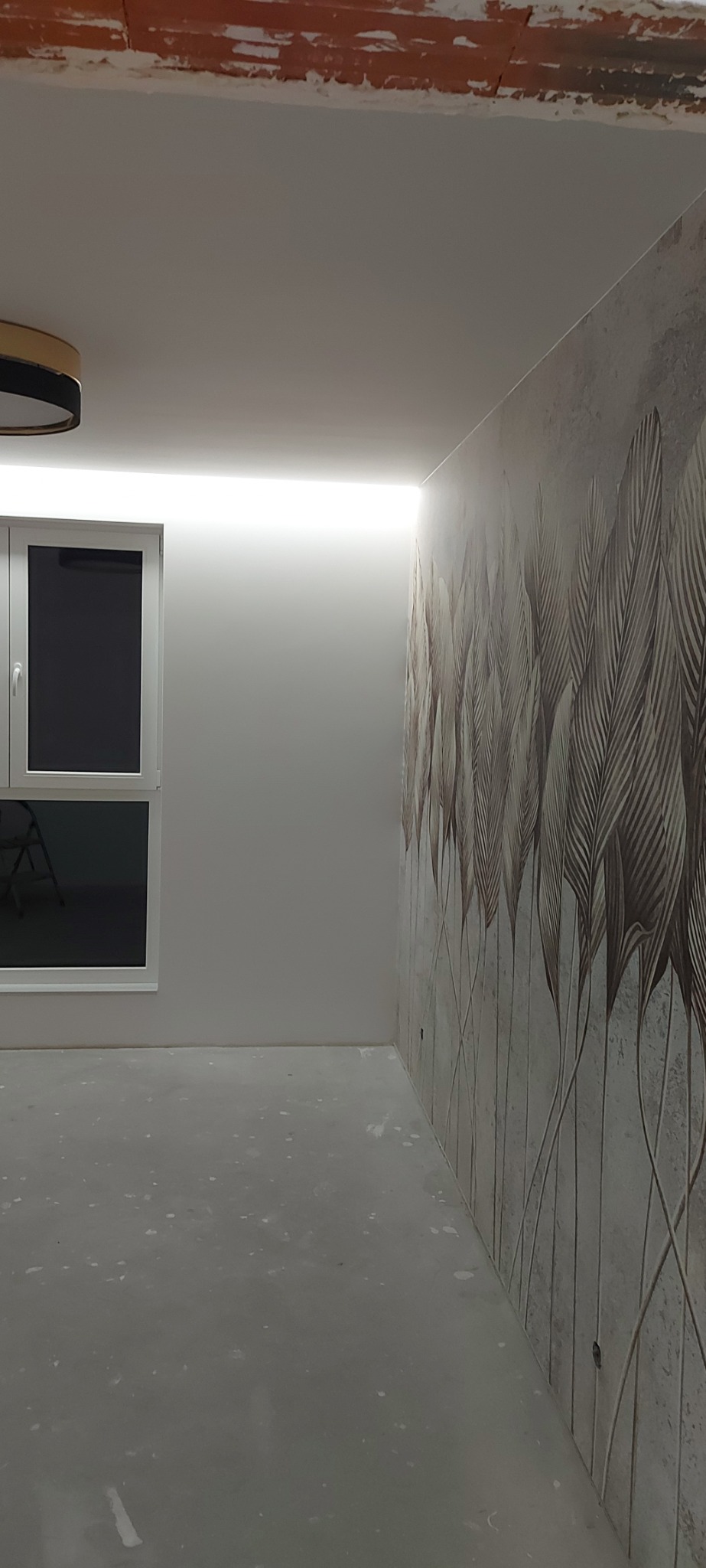 Pokój w trakcie prac wykończeniowych: szpachlowana podłoga, ściana z tapetą w motyw liści, sufit z oświetleniem LED, okno z widocznym krzesłem.