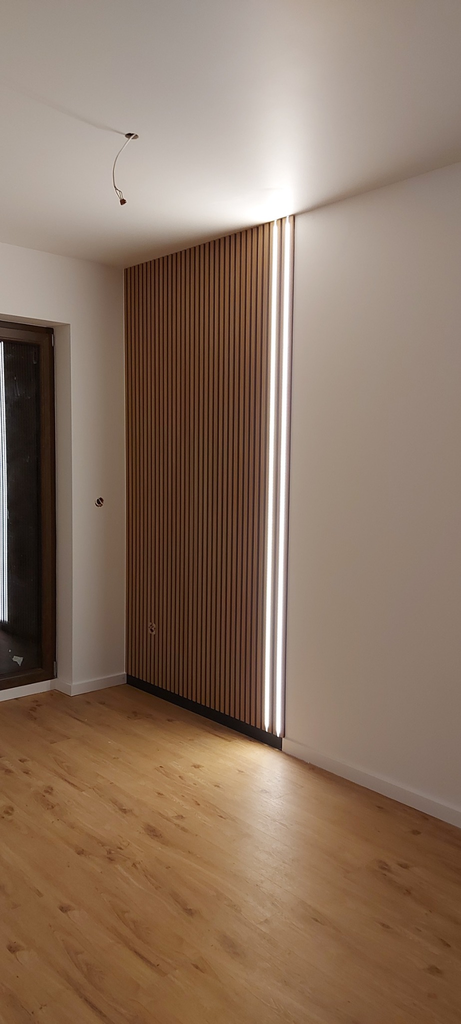 Nowoczesna ściana z drewnianych lameli z wbudowanym oświetleniem LED w jasnym pokoju z panelami podłogowymi, widoczne gniazdko elektryczne i wystające kable sufitowe.