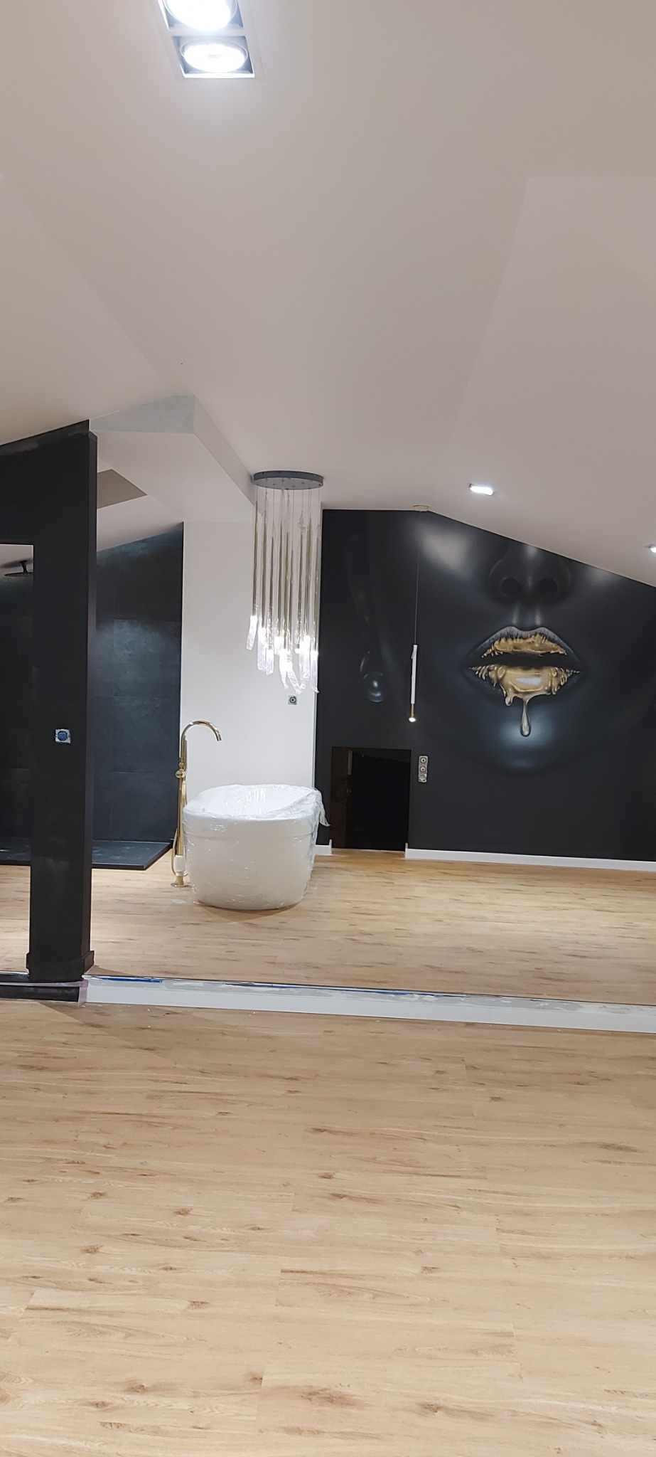 Nowoczesna łazienka z drewnopodobną podłogą i czarną ścianą z grafiką ust, wanna owinięta folią ochronną, wisząca lampa z kryształkami.