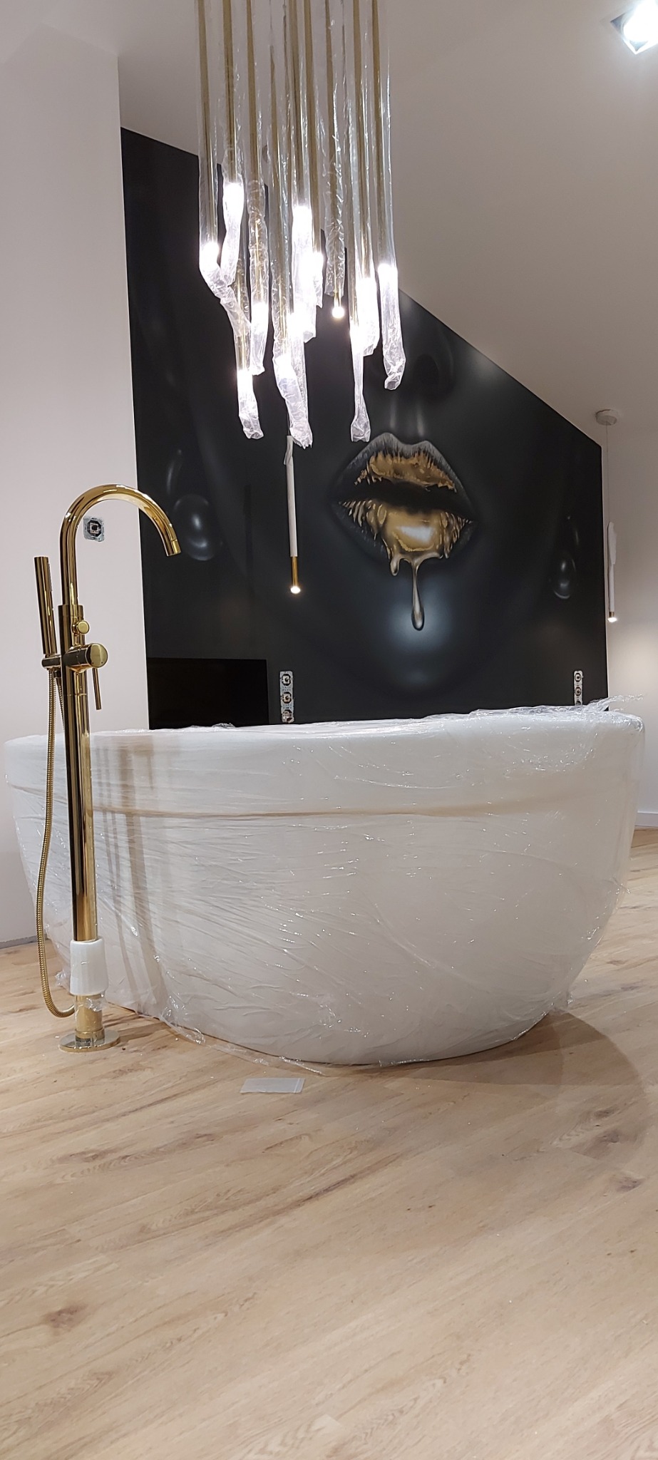 Nowoczesna łazienka z wolnostojącą wanną owiniętą folią ochronną, złotą baterią podłogową i designerskim żyrandolem nad wanną, na ścianie grafika przedstawiająca twarz kobiety.