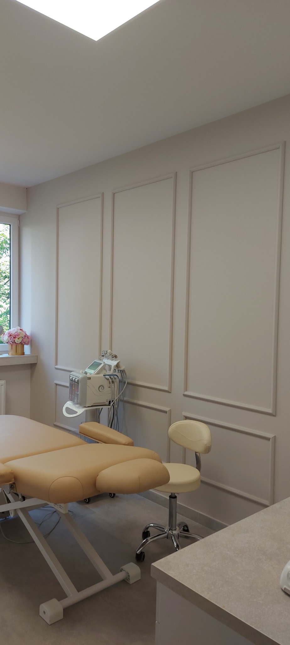 Jasne wnętrze gabinetu kosmetycznego z beżowym łóżkiem do zabiegów, krzesłem na kółkach i urządzeniem kosmetycznym na tle ściany z dekoracyjnymi listwami.