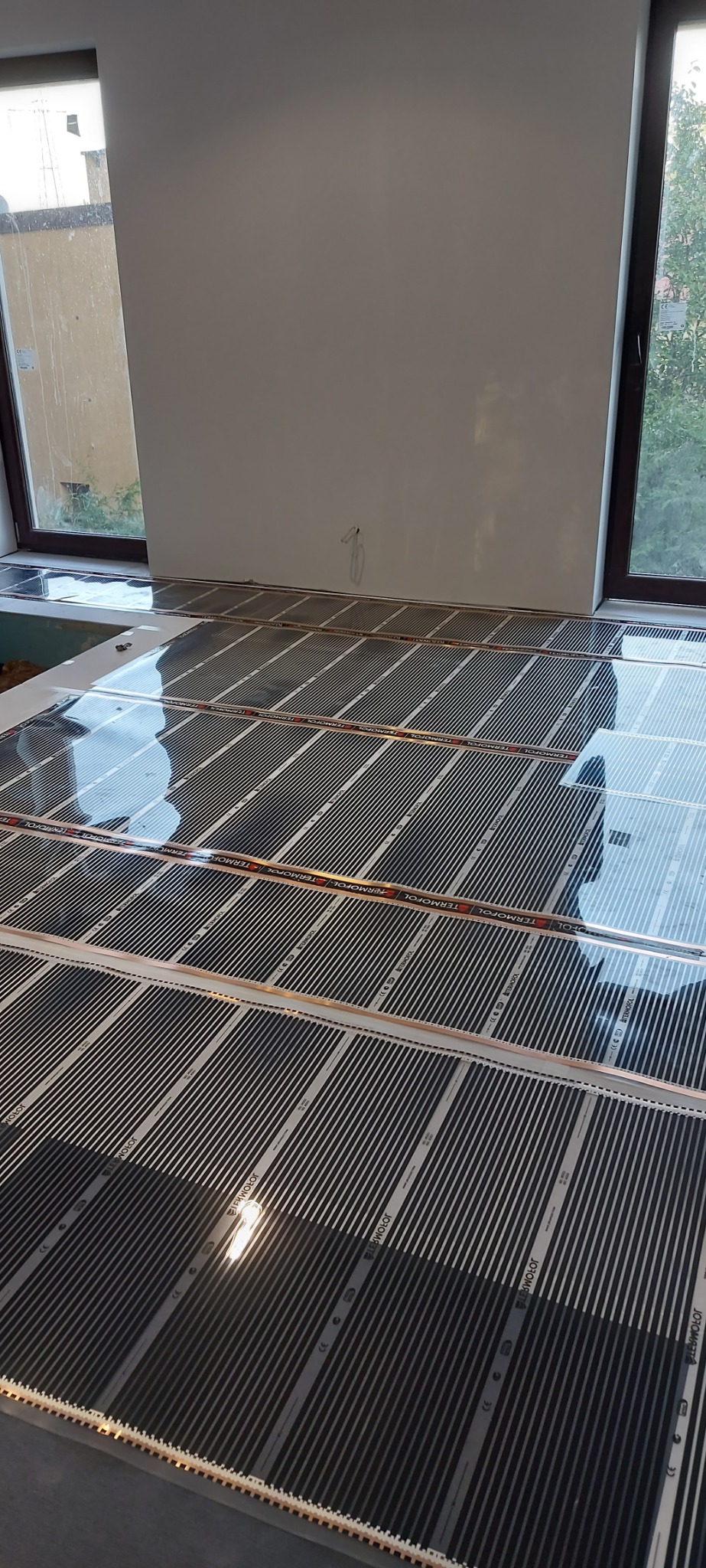 Rozłożona mata grzewcza pod panele podłogowe, widoczne okna i przygotowane ściany do dalszych prac wykończeniowych.