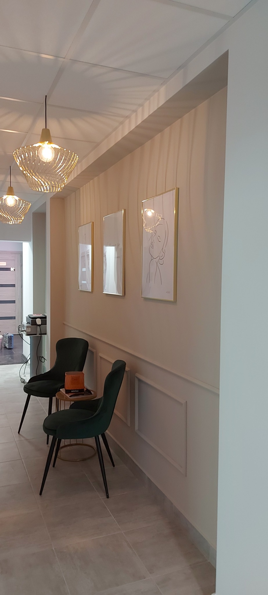 Jasne wnętrze poczekalni z kremowymi ścianami ozdobionymi sztukaterią i trzema obrazami w złotych ramkach, oświetlone dekoracyjnymi lampami wiszącymi, z dwoma ciemnozielonymi krzesłami i stolikiem...