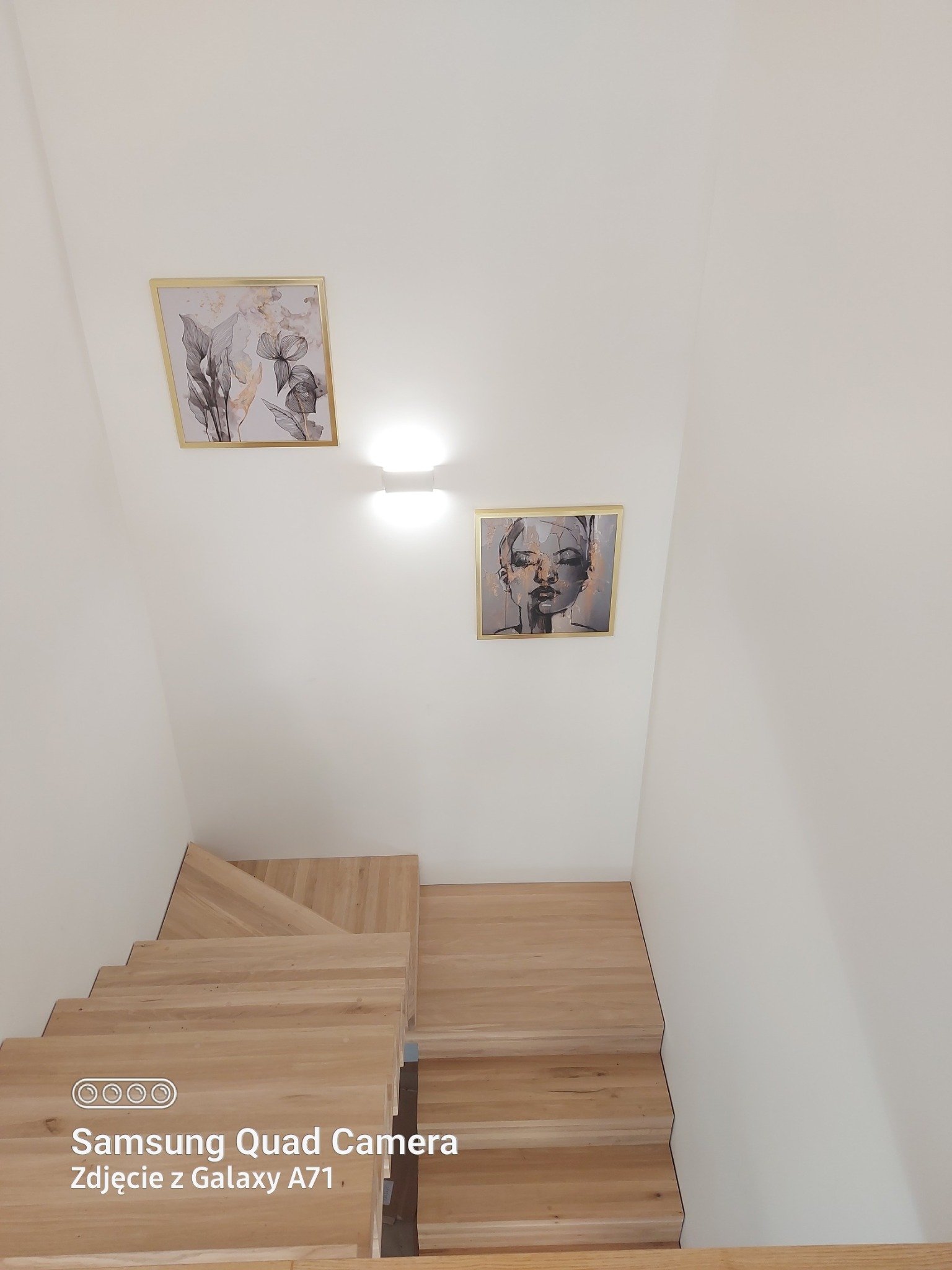 Nowe drewniane schody z jasnego drewna, widok z góry, białe ściany z dwoma obrazami w złotych ramach, oświetlenie sufitowe.