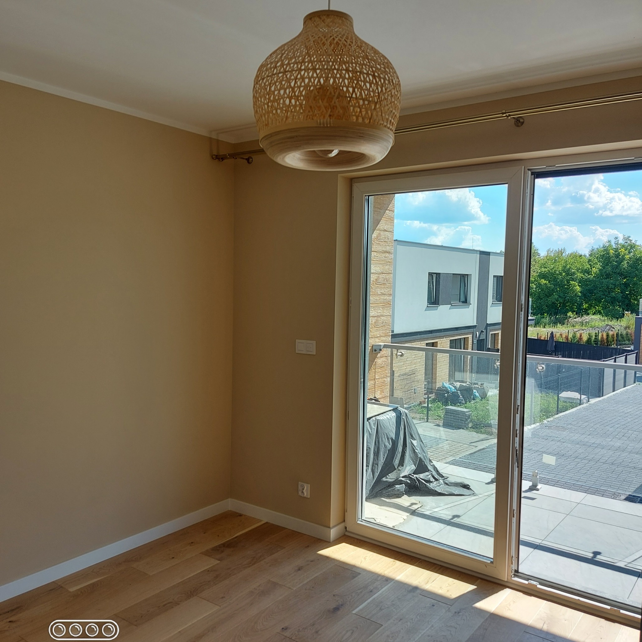 Beżowy pokój z drewnianą podłogą i plecioną lampą, widok na balkon przez przesuwne drzwi.