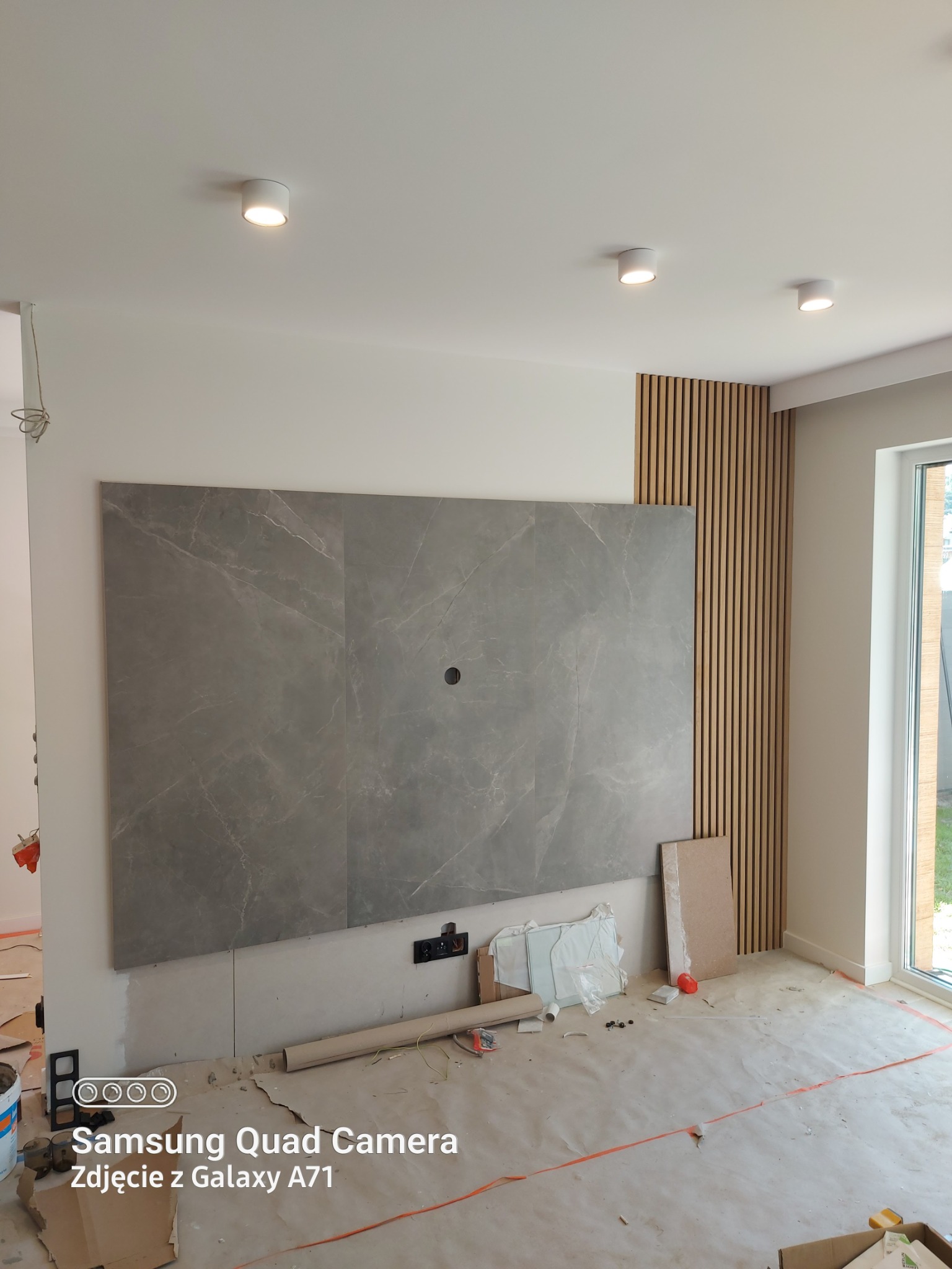 Ściana w trakcie wykańczania: szare płyty imitujące marmur, drewniane lamele pionowe, widoczne gniazdka elektryczne, podłoga zabezpieczona folią, sufit z oświetleniem punktowym.
