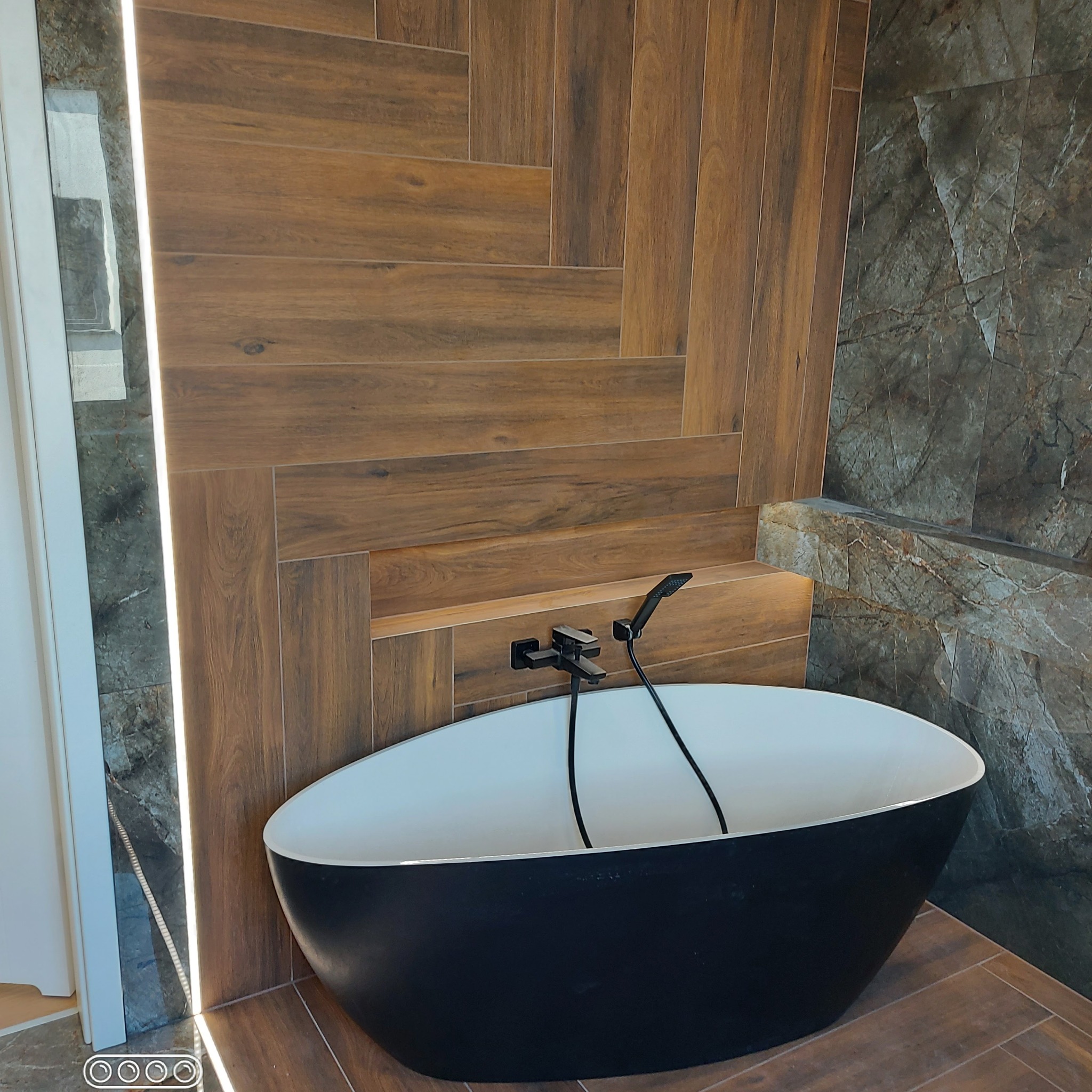Nowoczesna łazienka z wolnostojącą czarną wanną, drewnopodobne płytki ułożone w jodełkę na ścianie, marmurowe płytki po prawej stronie i podświetlenie LED po lewej.