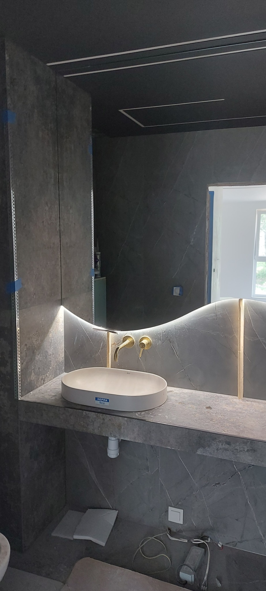 Szara łazienka w trakcie wykończenia, umywalka nablatowa z baterią w kolorze złotym, podświetlane lustro o falistym kształcie, widoczne narożniki i kable.