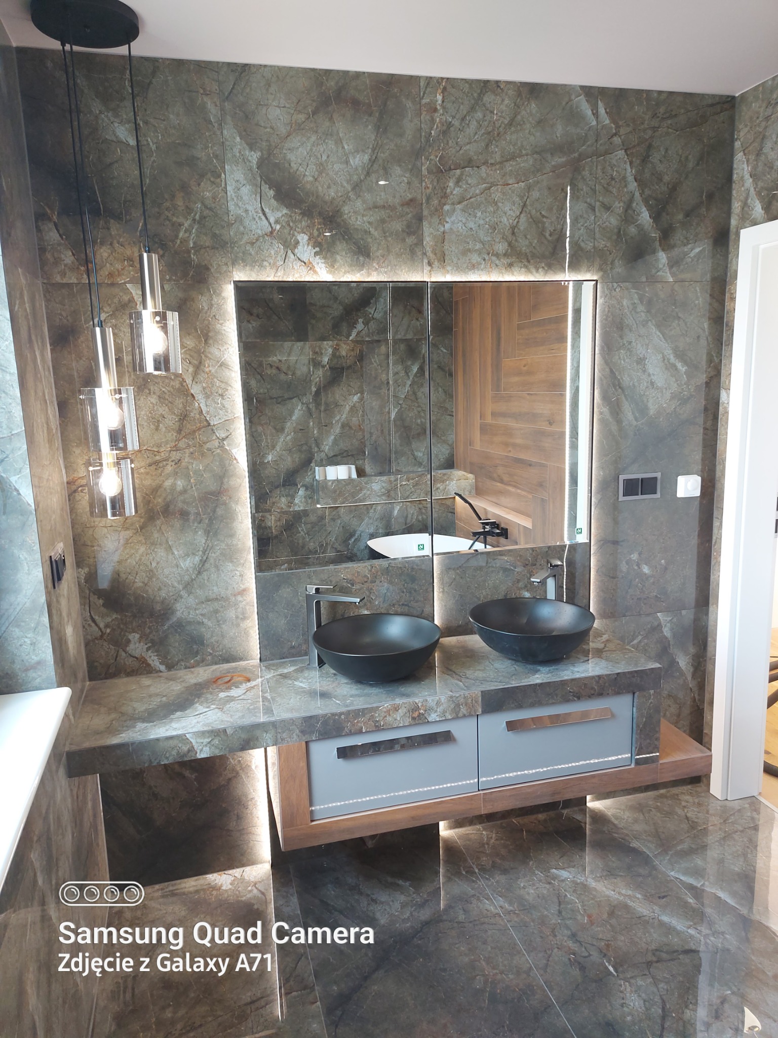 Nowoczesna łazienka z dwoma czarnymi umywalkami nablatowymi, podświetlanym lustrem i marmurowymi płytkami na ścianach i podłodze. Widoczne designerskie lampy wiszące i szafka pod umywalką...