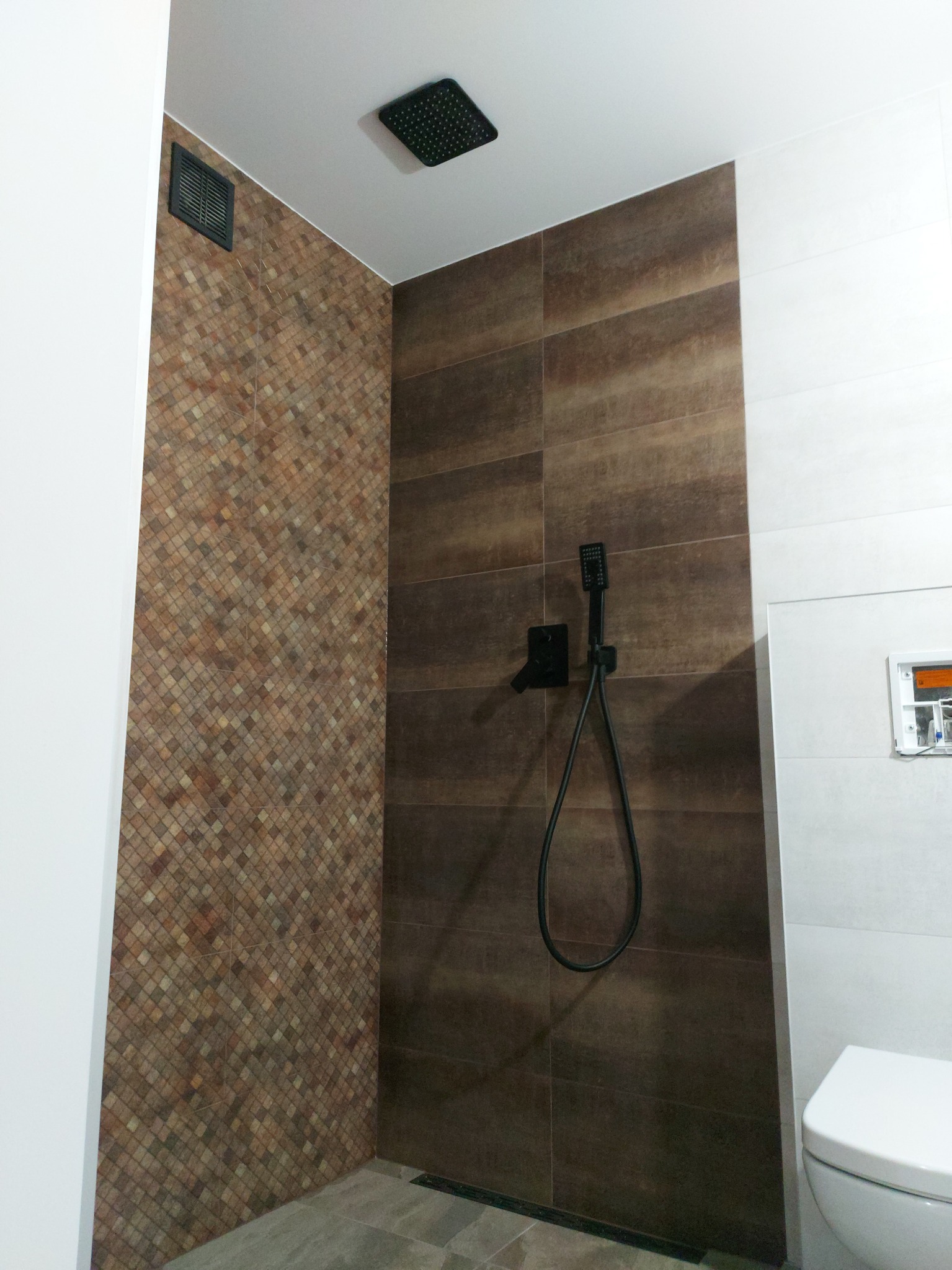 Nowoczesna kabina prysznicowa z brązowymi płytkami imitującymi drewno, mozaiką w odcieniach brązu i beżu oraz czarną armaturą, w tle widoczna toaleta.