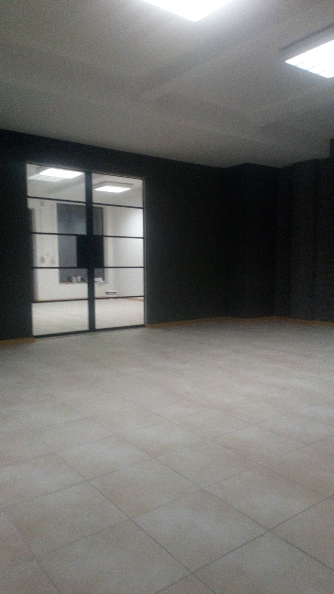 Pusty pokój z nowymi beżowymi płytkami podłogowymi ułożonymi w karo, ciemnymi ścianami i przeszklonymi drzwiami z czarnymi szprosami.