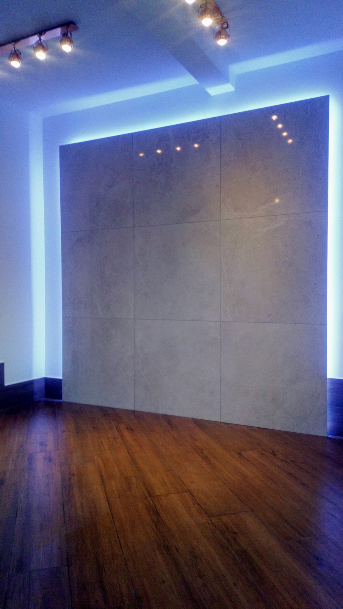 Ściana wyłożona dużymi płytami w odcieniach szarości, podświetlona niebieskim światłem LED, z parkietem na podłodze i reflektorami na suficie.
