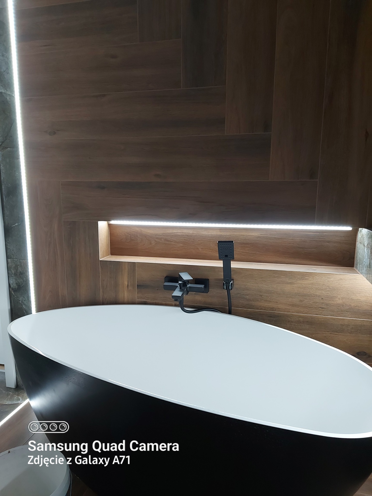 Nowoczesna łazienka z wolnostojącą czarną wanną, drewnopodobnymi płytkami na ścianie z podświetlaną półką i szarą ścianą z oświetleniem LED.