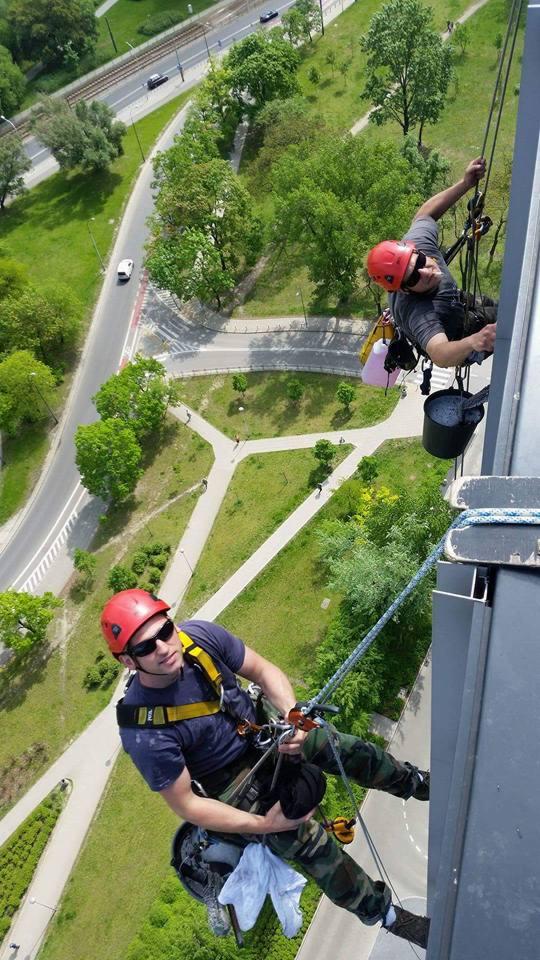 Dwóch alpinistów przemysłowych w czerwonych kaskach, zawieszonych na linach, czyści elewację wysokiego budynku z widokiem na park i ulicę.
