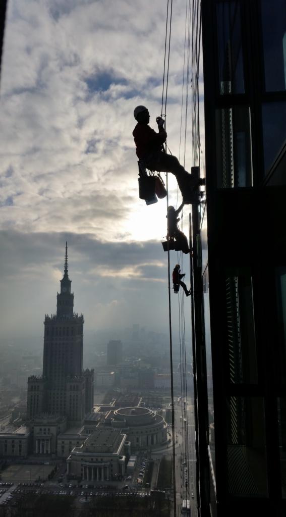 Silhouettes of three window cleaners suspended on ropes, working on a high-rise building with the Palace of Culture and Science visible in the background under a cloudy sky.