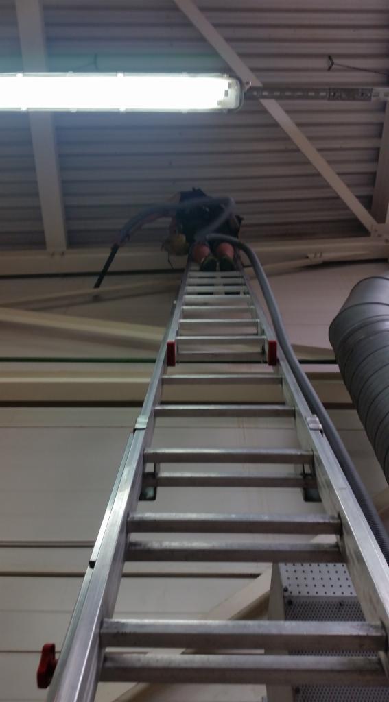 Pracownik na drabinie, czyszczący sufit hali przemysłowej za pomocą odkurzacza przemysłowego z długim wężem, widok z dołu na oświetlenie i konstrukcję dachu.