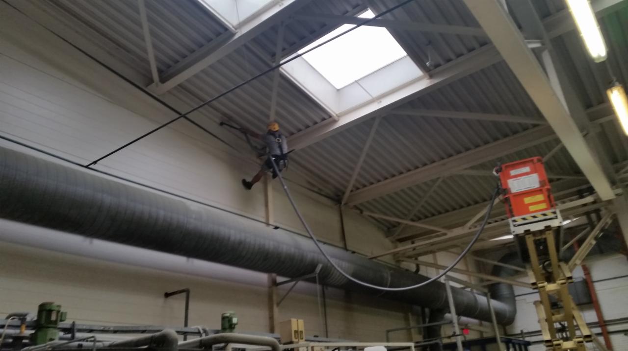 Pracownik w kasku i uprzęży, wiszący na linie, czyści rury wentylacyjne pod sufitem hali przemysłowej w Warszawie. W tle widoczny podnośnik z pomarańczową skrzynką.