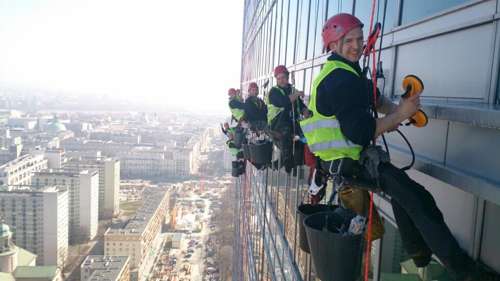 Grupa alpinistów przemysłowych w kaskach i kamizelkach odblaskowych myje okna wieżowca w Warszawie, widok na miasto w tle.