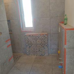 Krzyś - Łazienka w trakcie wykańczania: szare, kwadratowe płytki na ścianach i podłodze, okno z pomarańczową taśmą, zabudowa WC z dekoracyjnymi płytkami i butelka wody na górze, widoczne elementy...