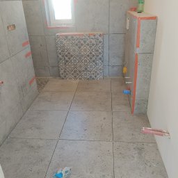 Krzyś - Szare płytki podłogowe i ścienne w trakcie montażu w łazience, widoczne krzyżyki dystansowe i elementy instalacji hydraulicznej, butelka z płynem na obudowie stelaża podtynkowego.