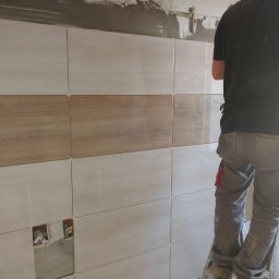 Krzyś - Układanie płytek imitujących drewno na ścianie, widoczne krzyżyki dystansowe i otwory na instalacje wodne, pracownik na drabinie w tle.